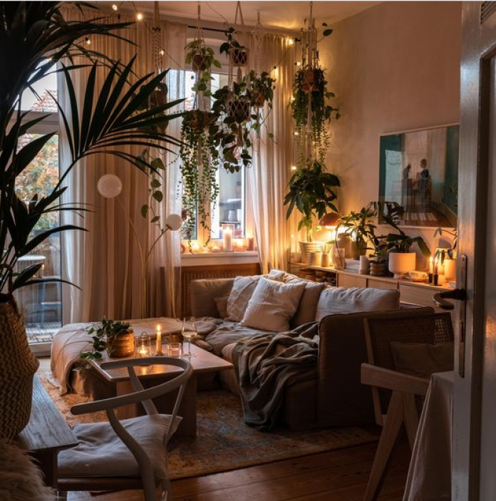 menajare living boem și eclectic, cu o canapea confortabilă, plante suspendate din tavan în fața ferestrei, ghirlande luminoase calde și lumânări aprinse.
